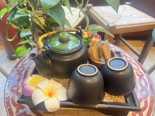 Présence d'un ensemble de deux tasses et d'une jolie théière sur un plateau. Deux biscuits accompagne le toux avec deux fleurs de frangipanier.